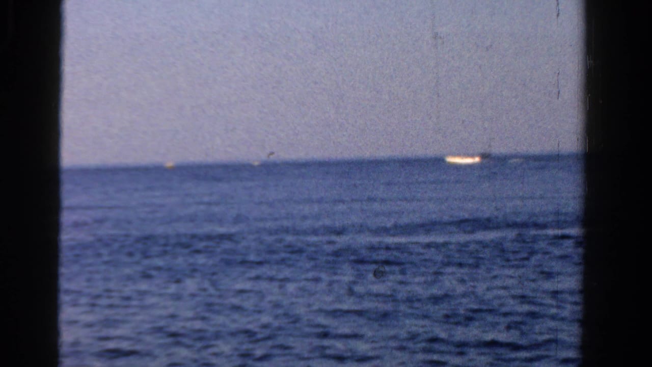 Open water and pale sky seen through dark vertical boat framing, with a small bright vessel near the horizon.