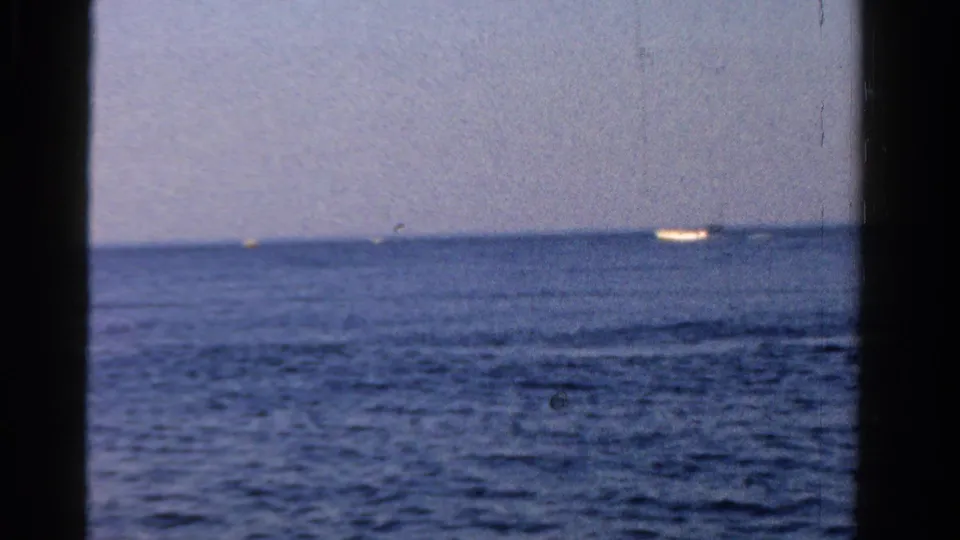 1958 Ontario home-movie frame showing rippled open water, a pale sky, dark boat framing, and a distant vessel on the horizon.
