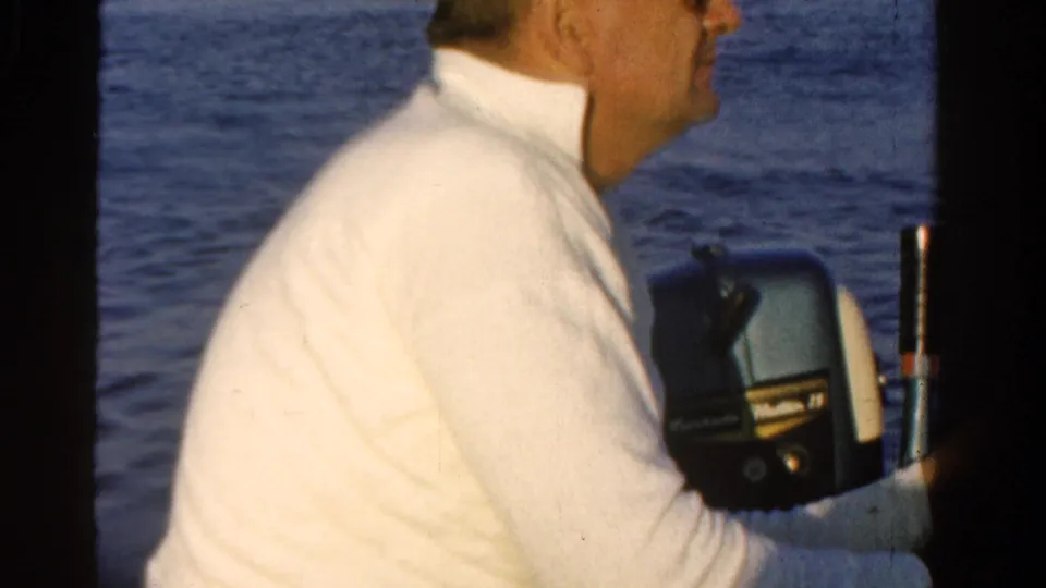 Adult man in a white sweater sits at the motor area of a boat on open water in 1958 Ontario home-movie footage.