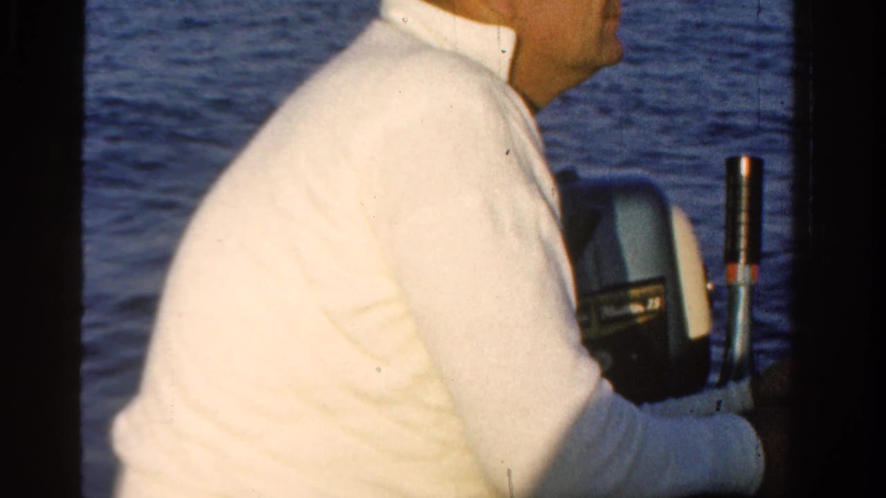 Man in a white shirt sits beside an outboard motor on blue water.