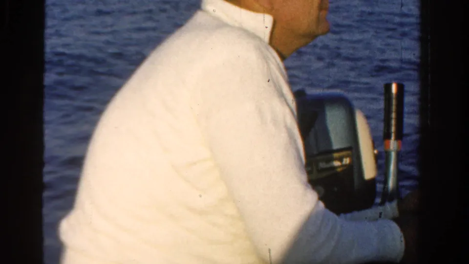 In this 1958 Ontario 8mm frame, a man in a white shirt sits at the side of a small boat near an outboard motor on calm blue water.
