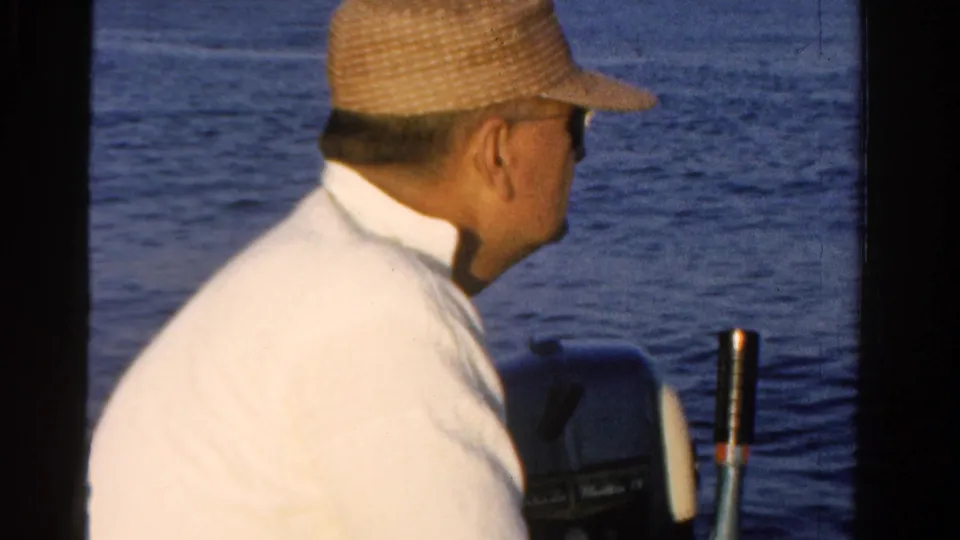 A 1958 Ontario boating frame shows a man in a straw hat and white shirt looking across the water while seated near the motor.