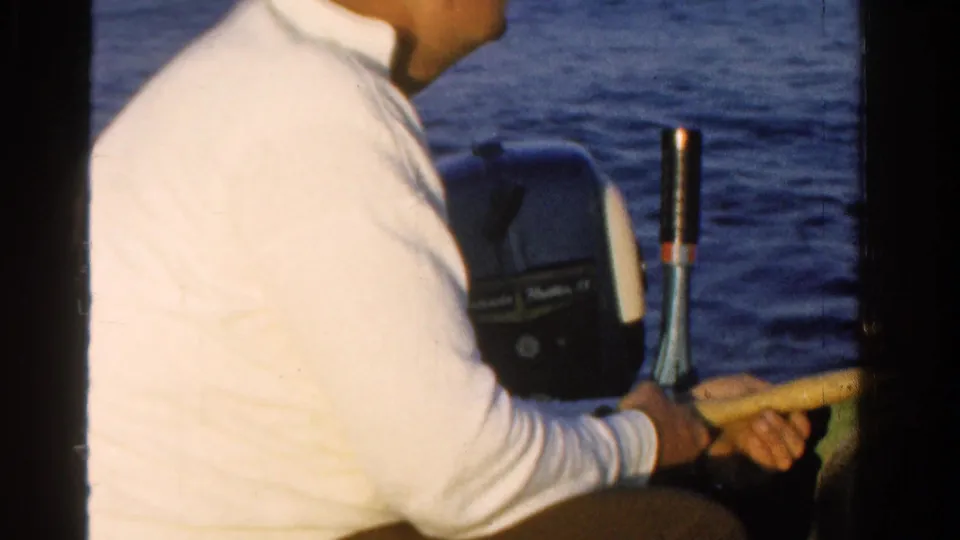 The home-movie frame shows the man holding a boat control or tiller near the outboard motor as calm water fills the background.