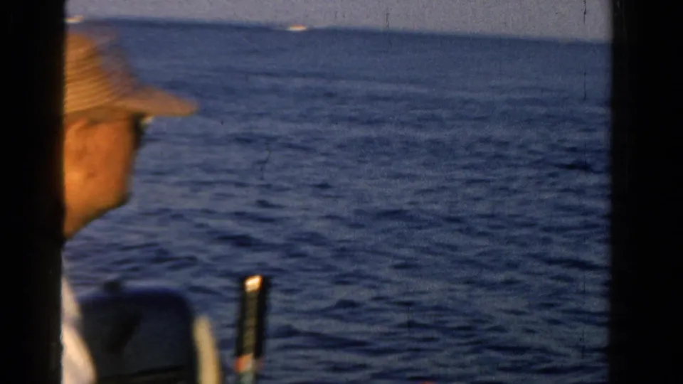 1958 Ontario home-movie frame with a partial close view of a hat-wearing man beside open water and dark frame edges.