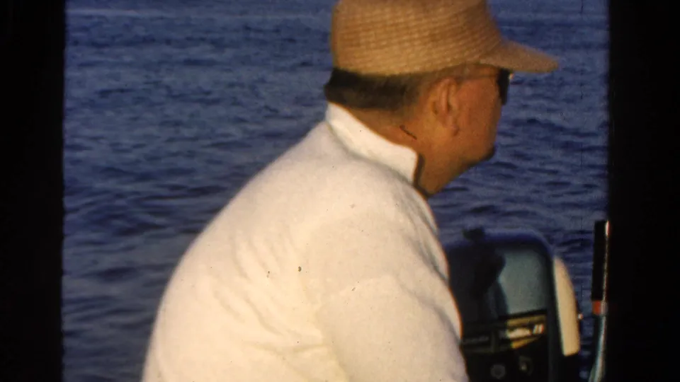 A man in a straw hat sits near the boat motor, facing away toward the water in 1958 Ontario footage.
