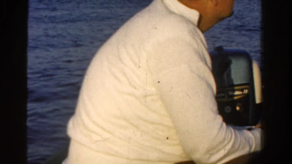 1958 Ontario home-movie frame of a man in a white shirt seated by dark boat framing as blue water fills the background.
