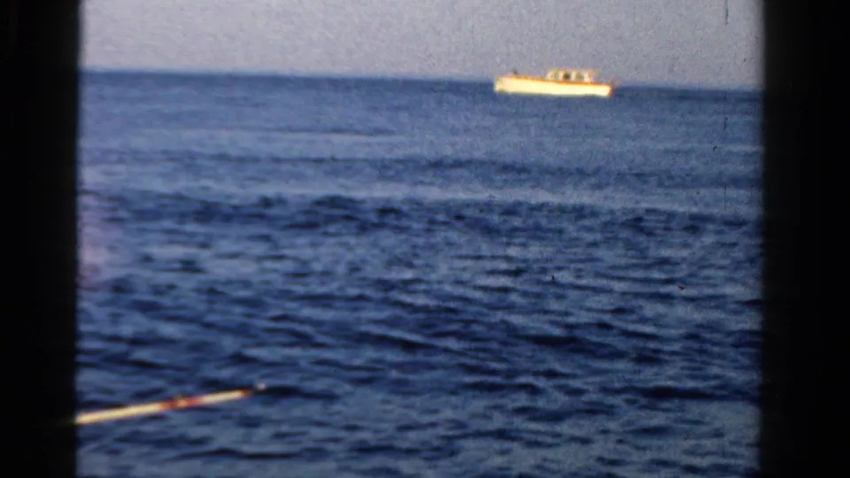 1958 Ontario 8mm frame showing a small motorboat far across calm blue water, seen through dark boat or window edges.