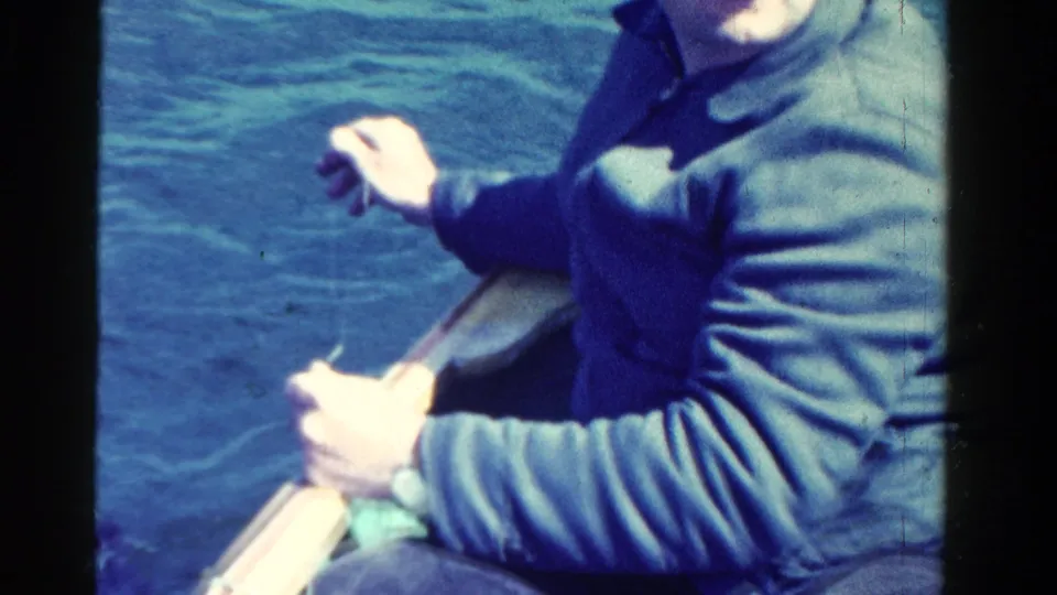 Close 8mm frame of a man in a blue jacket seated in a rowboat on an Ontario lake, based on supplied 1958 metadata.