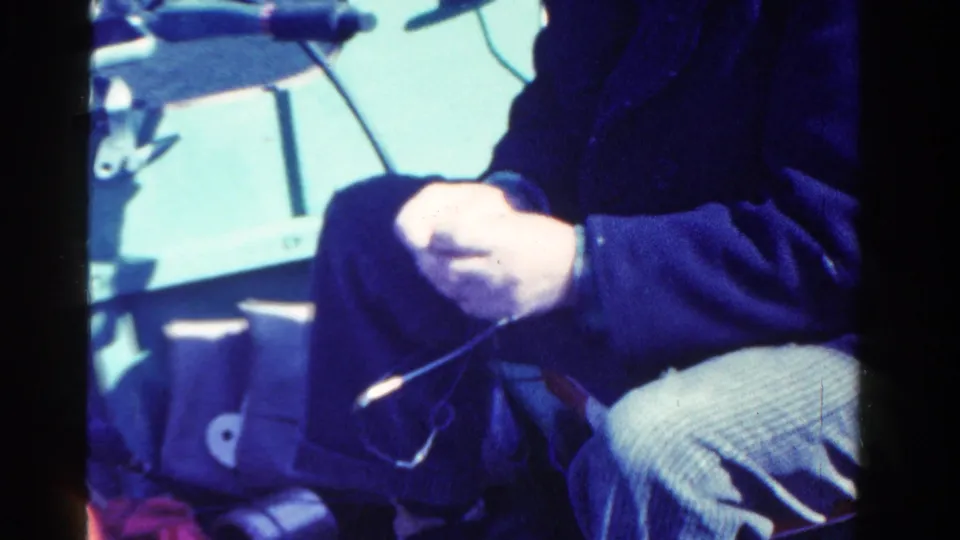 In 1958 Ontario home-movie footage, a seated fisherman in a dark coat works with fishing line beside a tackle box on a boat.