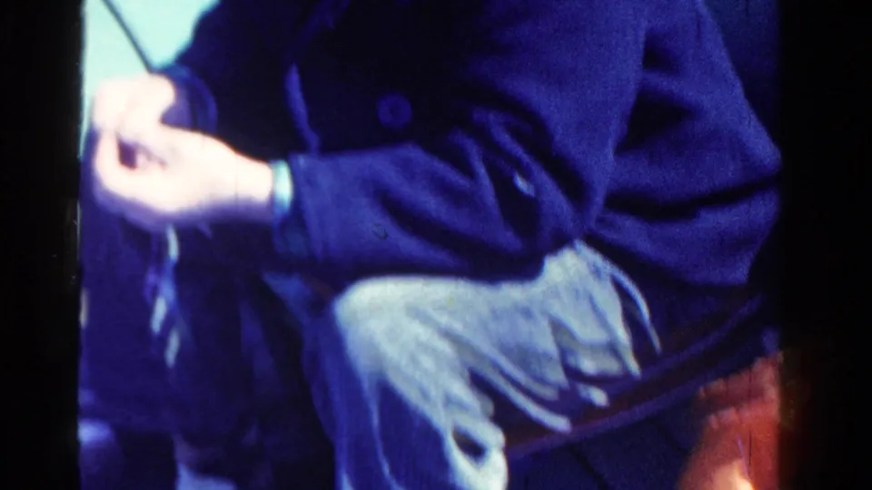A seated fisherman in a dark coat and corduroy pants works with line or bait on a boat in the 1958 Ontario home movie.