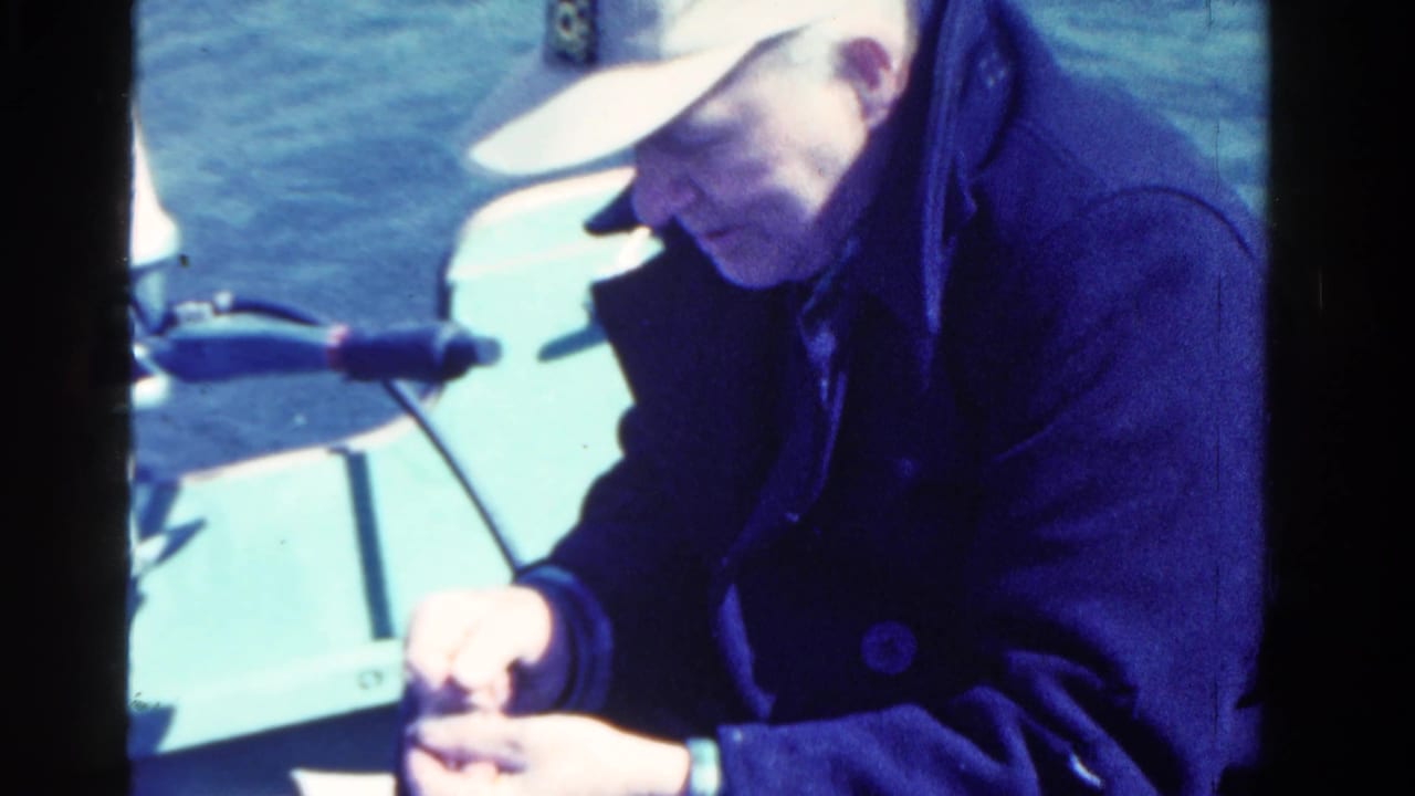 Man in a cap and dark coat handling a small object aboard a boat.