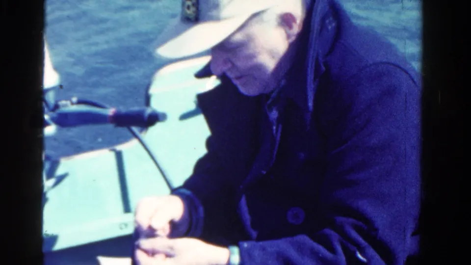 In 1958 Ontario home-movie footage, a man in a cap and dark coat looks down while handling a small object on a boat.