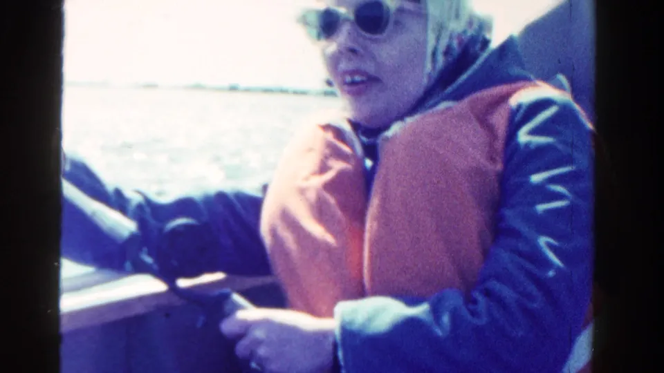 A woman in sunglasses, a headscarf, and an orange life vest smiles while riding in a boat in 1958 Ontario footage.