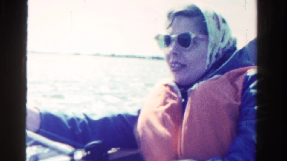 In Ontario, Canada in 1958, a woman in sunglasses, headscarf, and orange life vest sits on a boat with water behind her.
