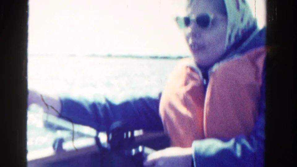 The woman remains seated in the boat, wearing sunglasses, a headscarf, and an orange life vest against a bright water background.