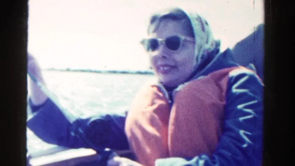 In Ontario, Canada, 1958, a woman in a headscarf, sunglasses, blue jacket, and red life vest fishes from a small boat.