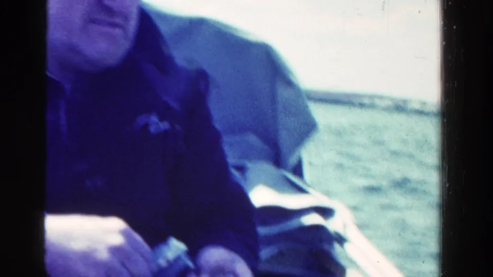 1958 Ontario home-movie frame showing a seated man in dark clothing in a boat, with water and boat seating behind him.