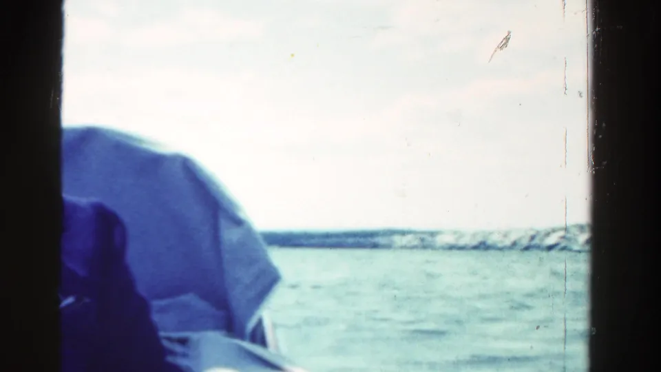 Home-movie frame from Ontario showing part of the boat interior, nearby water, and a low shoreline in the distance.