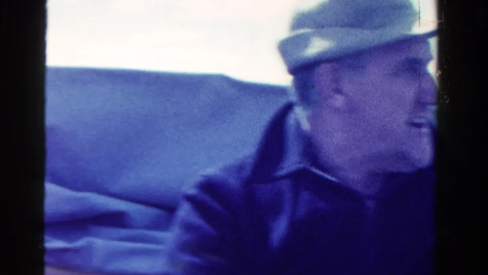 1958 Ontario home-movie frame showing a man in a brimmed hat and dark jacket seated in a small boat.
