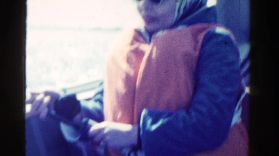 A woman aboard a boat in 1958 Ontario footage holds fishing gear, with water and bright sky behind her.