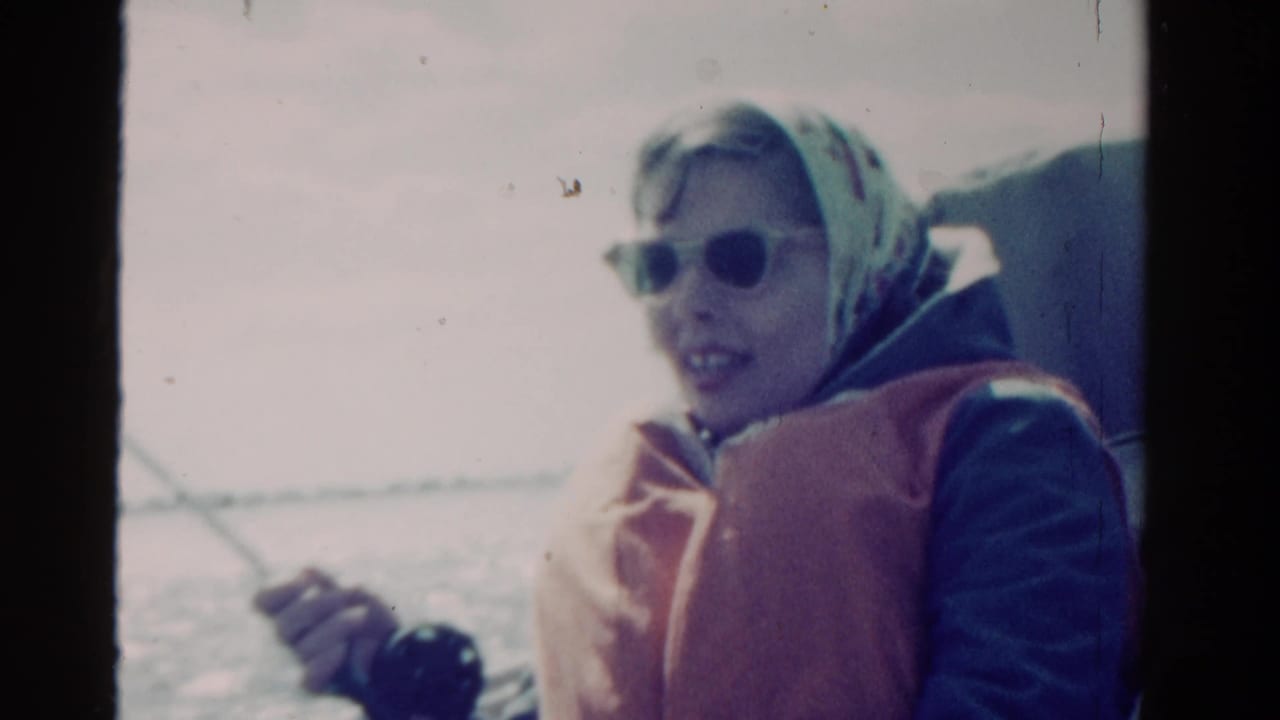 1958 Ontario home-movie frame of a woman in sunglasses, headscarf, and life vest holding a fishing rod on open water.
