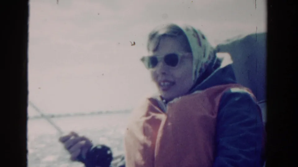 1958 Ontario home-movie frame of a woman in sunglasses, headscarf, and life vest holding a fishing rod on open water.