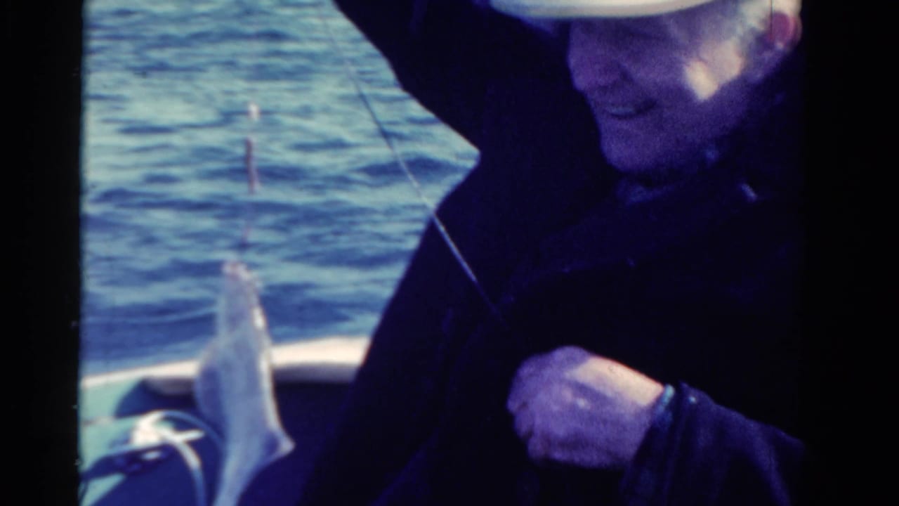 1958 Ontario home-movie frame of a seated adult man in a small boat holding a caught fish on a line over blue water.