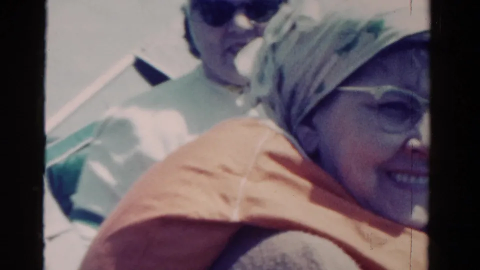 Older woman smiles toward the camera in a headscarf, sunglasses, and orange life vest during a 1958 Ontario boat trip.