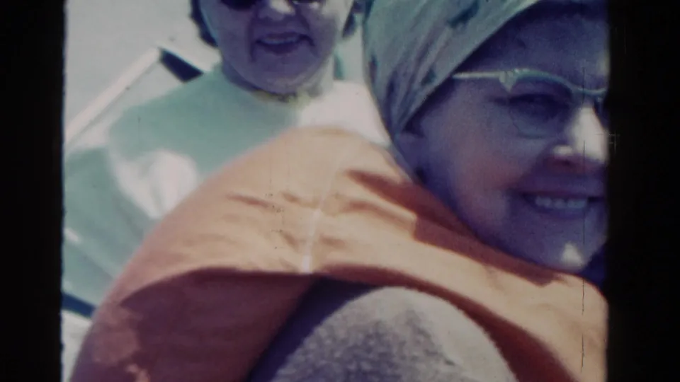 Close home-movie view of smiling boat passengers in Ontario, with a headscarf, glasses, and orange life vest visible.