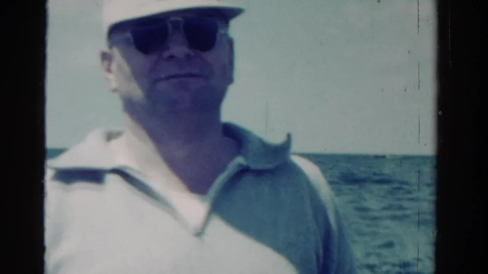 Man in a cap and sunglasses stands on the boat deck with open water behind him in the supplied 1958 Ontario footage.
