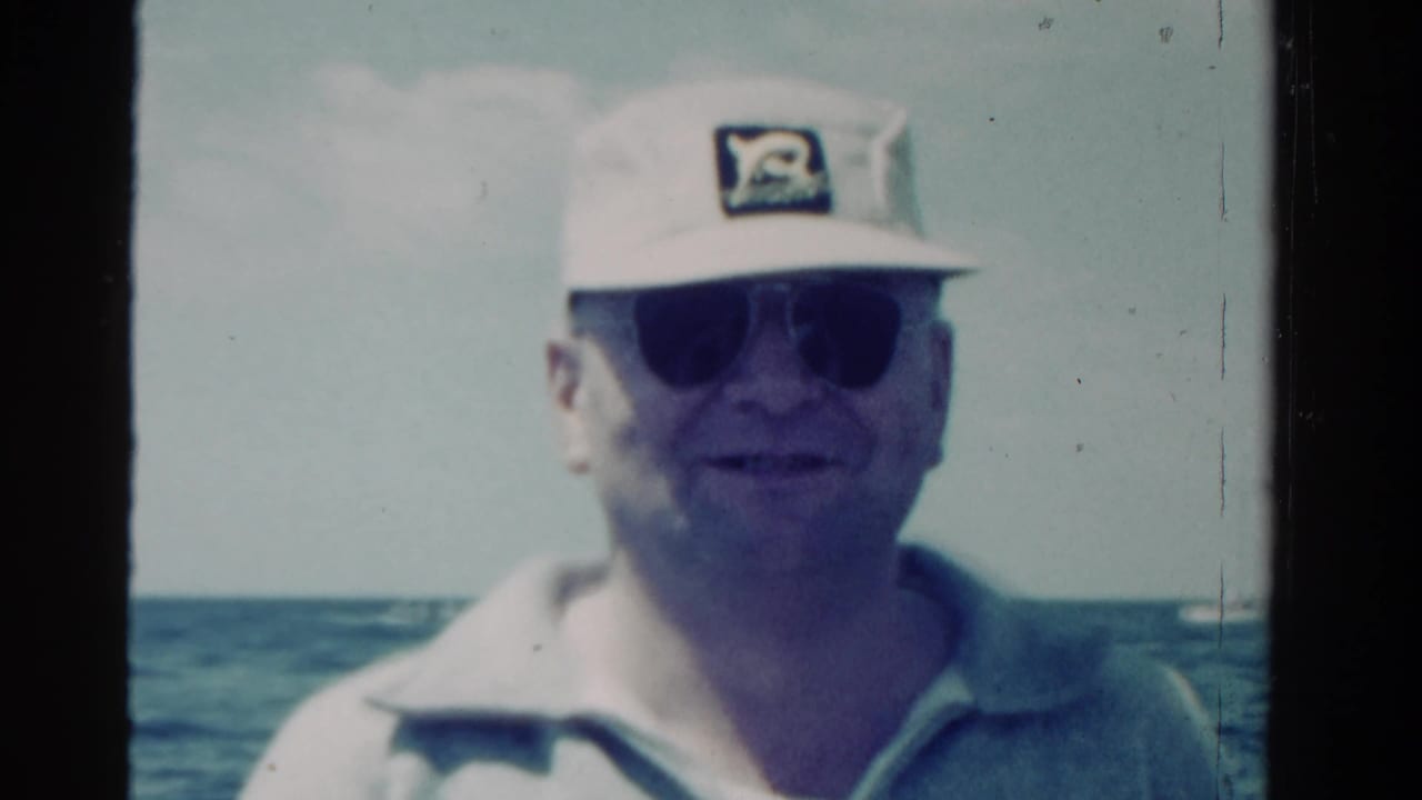 Man in cap and sunglasses smiles slightly on a boat with horizon behind him.