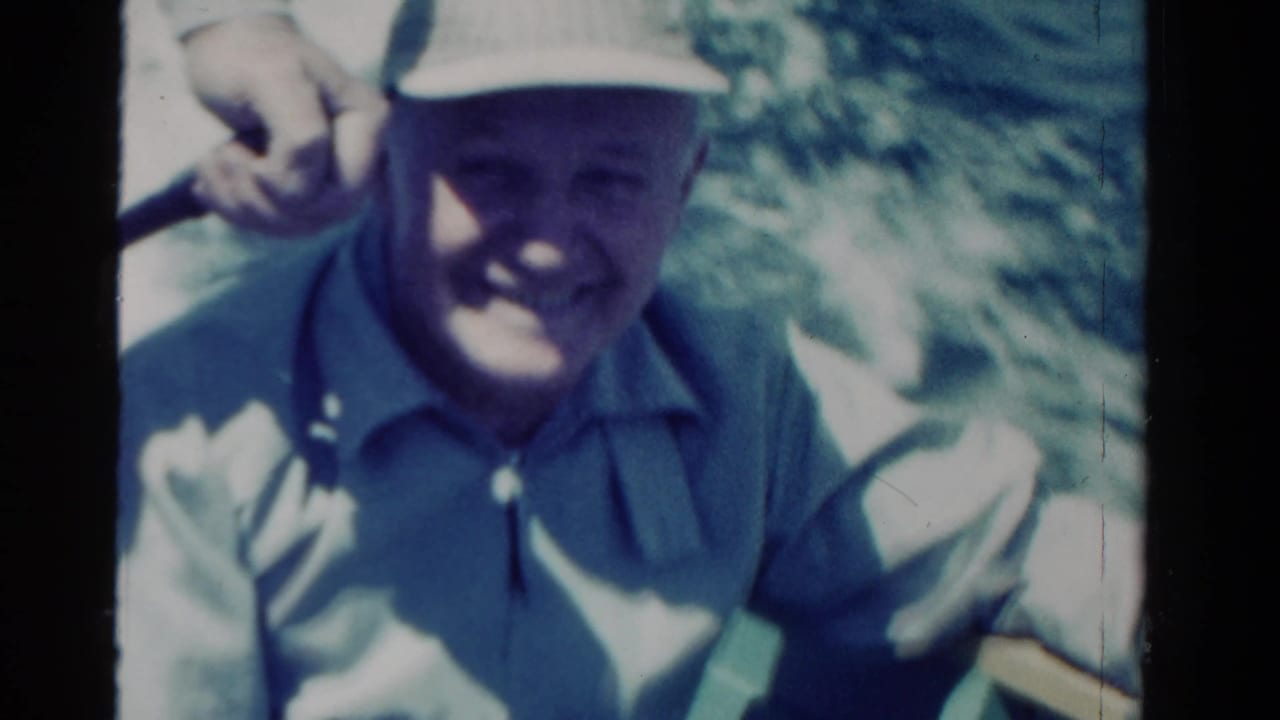 Man in a white cap raises his hand while seated on a boat.