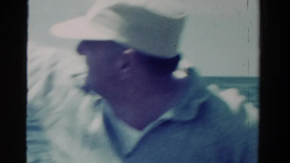 The man turns sideways in the boat, with his white cap and pale shirt visible in soft, faded 8mm color.