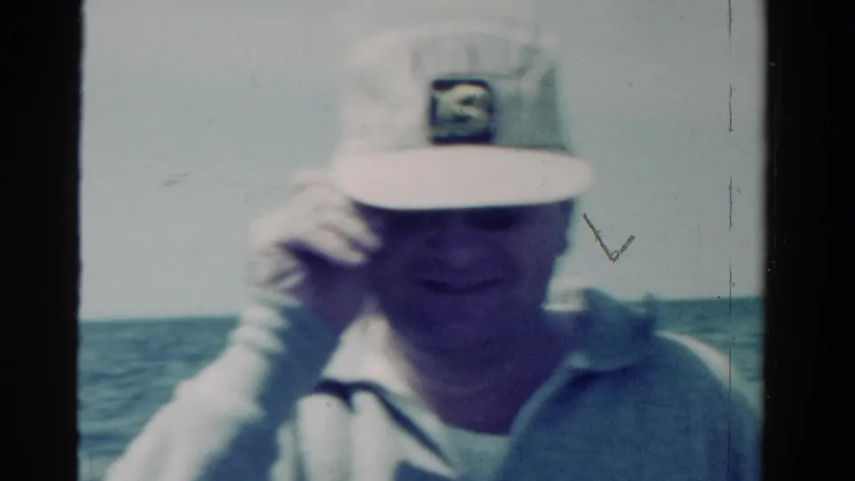 The man adjusts his white cap while seated on the boat, with open water behind him in the 1958 Ontario footage.