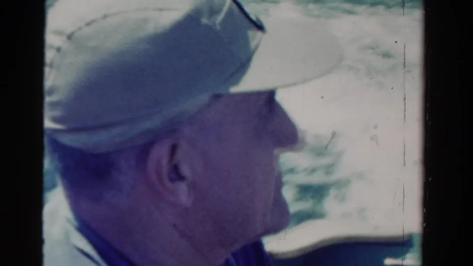 Side view of the man seated on a boat in a light cap, with bright water behind him in 1958 Ontario home-movie footage.