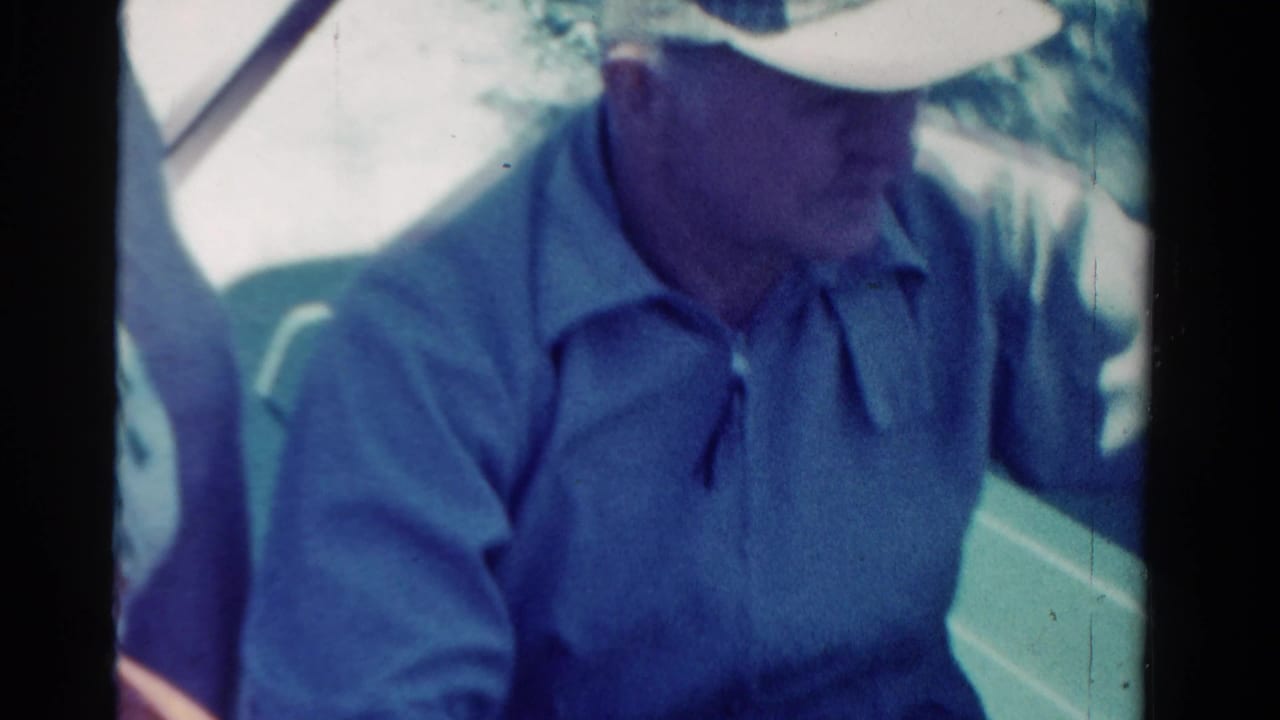 1958 Ontario home-movie frame of an adult man in a blue shirt and light cap seated by a car window during travel.