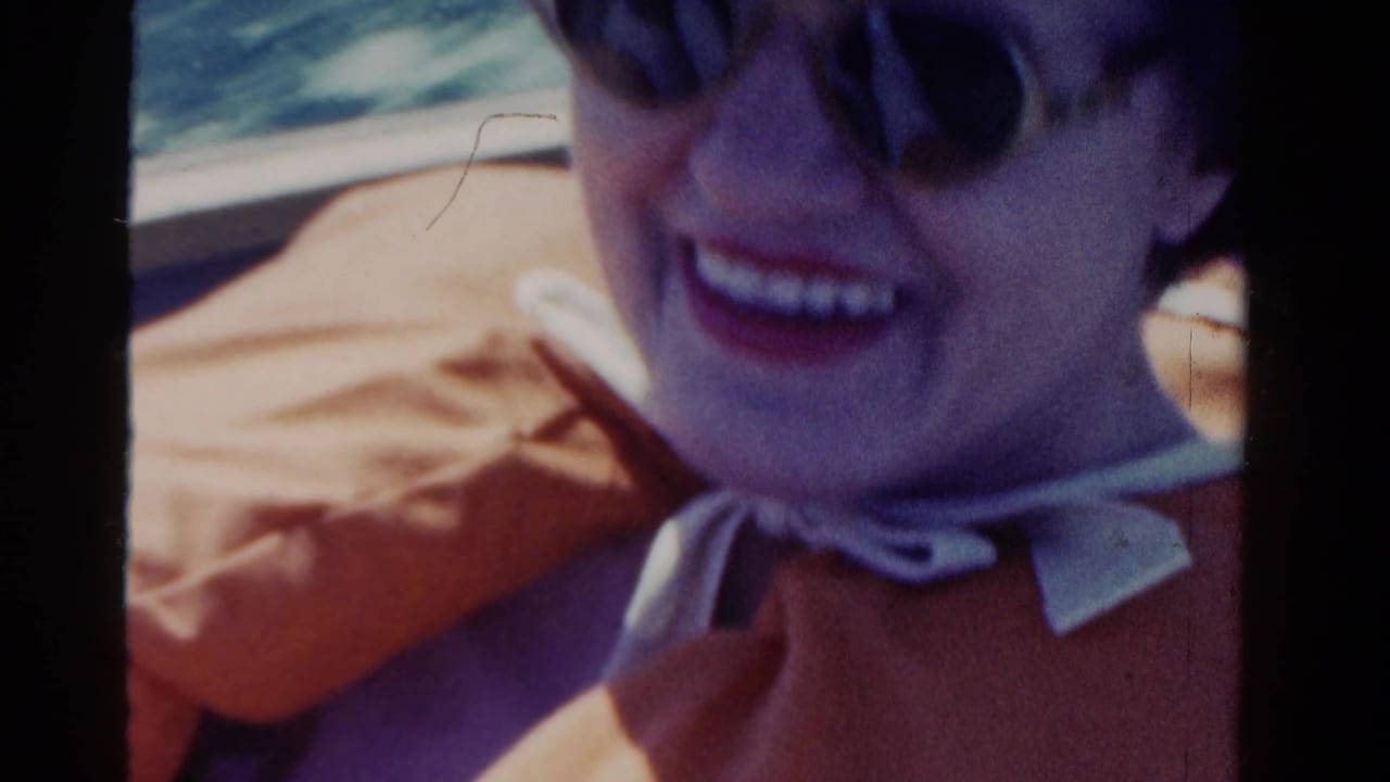 1958 Ontario 8mm frame showing a smiling woman in sunglasses, a pale hat, and orange clothing during a sunny vehicle ride.
