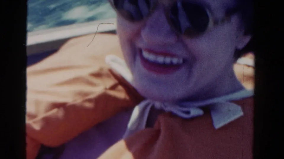 1958 Ontario 8mm frame showing a smiling woman in sunglasses, a pale hat, and orange clothing during a sunny vehicle ride.