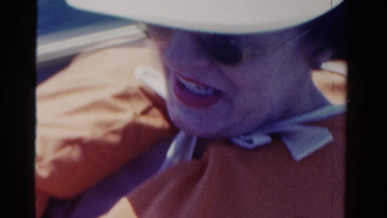 1958 Ontario home-movie frame of a woman in a white hat and dark sunglasses smiling from an open vehicle.