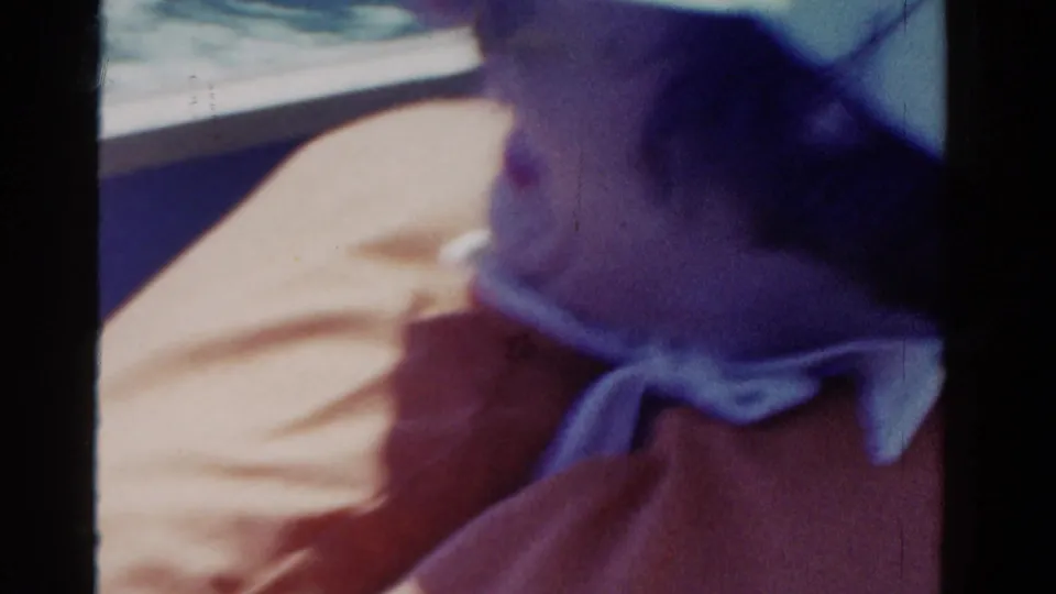 Soft 8mm frame from 1958 Ontario showing the woman turning in the vehicle, with sunglasses, bow collar, and orange clothing visible.