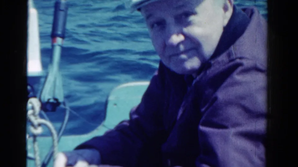 1958 Ontario 8mm frame of a man in a dark jacket seated on a small boat, with blue water, rope, and boat railings behind him.