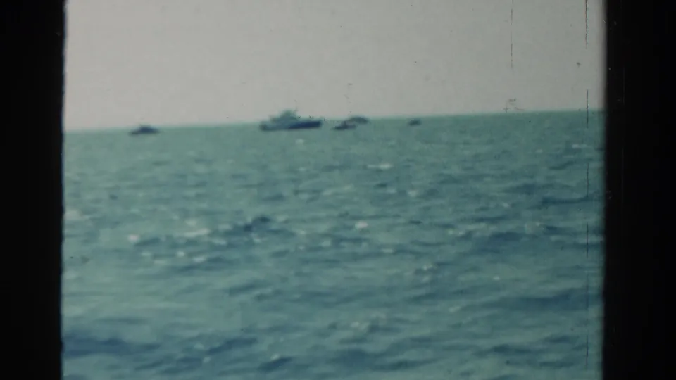 1958 home-movie view over blue-green open water, with several small boats or dark shapes near the distant horizon.