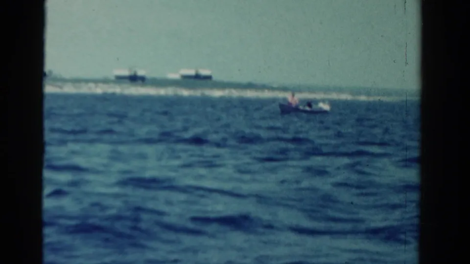 1958 Ontario 8mm frame showing a small flag-bearing boat on open water with distant vessels and wake near the horizon.