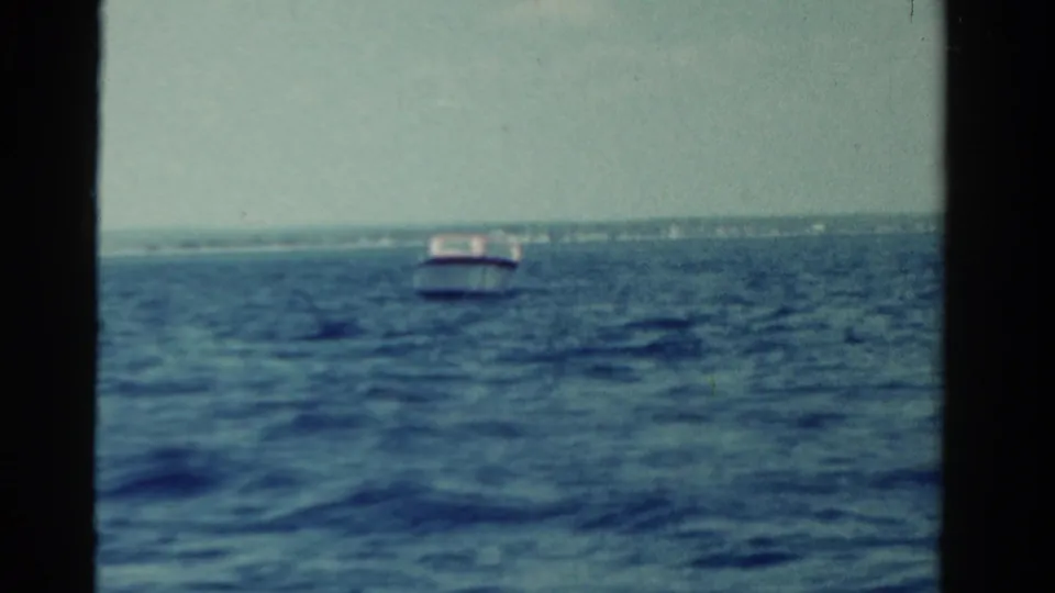 1958 Ontario home-movie frame with a small boat ahead on blue water and a low shoreline stretching along the horizon.