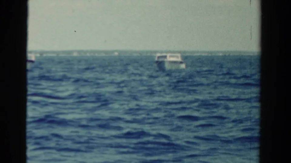 1958 Ontario 8mm frame showing a small boat on rippled water, filmed from another vessel with a low shore in the background.