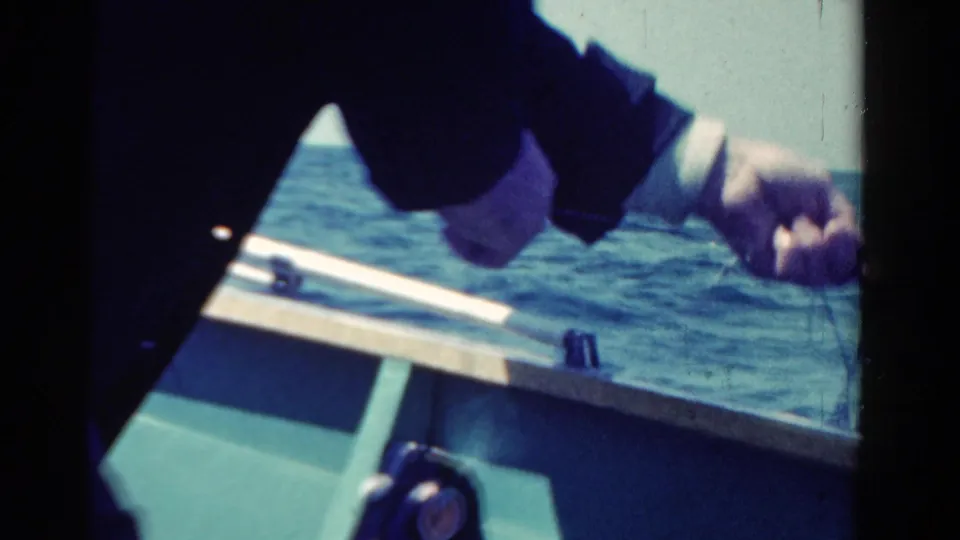 1958 8mm frame of a person bent over the boat side, with fishing line and blue water filling the background.