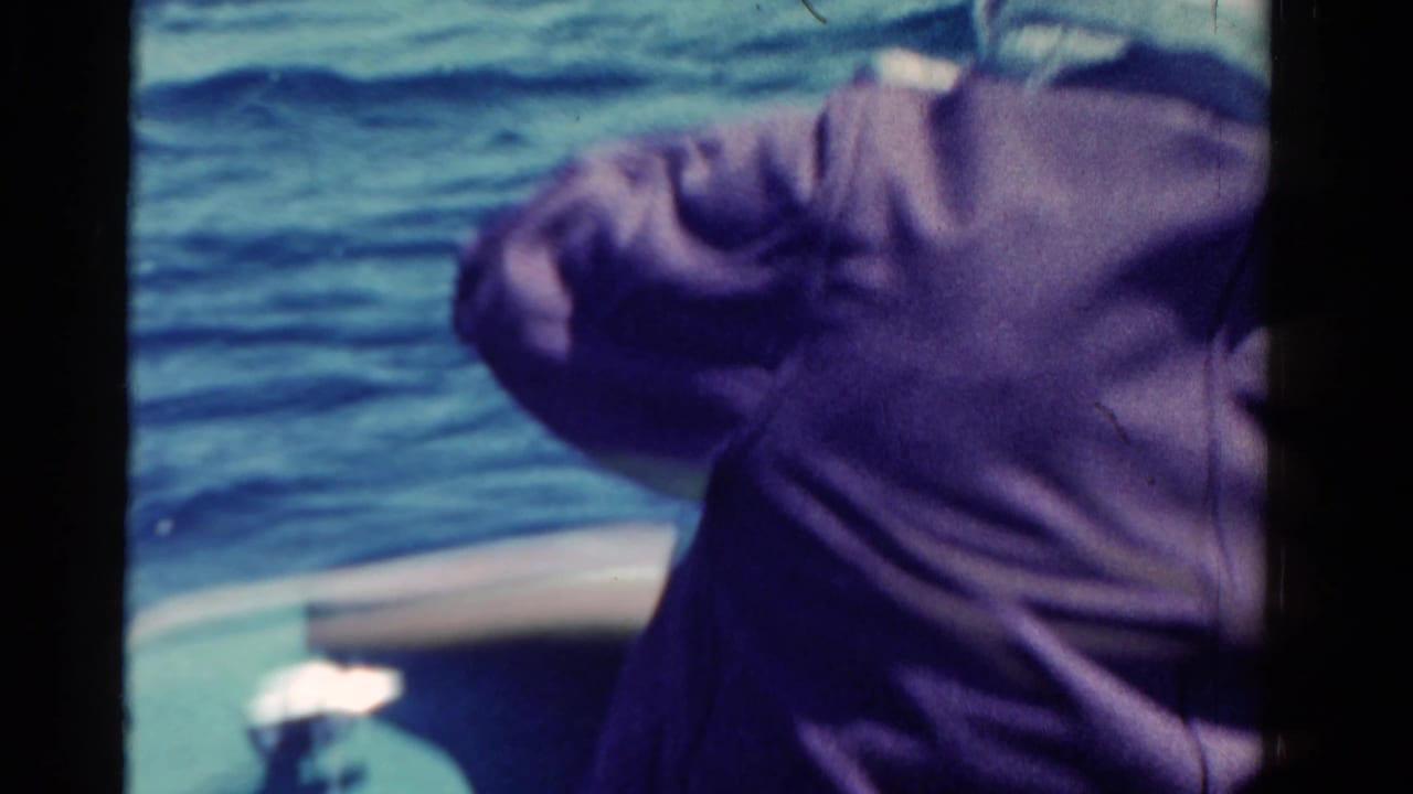 Man in a dark jacket leans forward on a small boat beside blue water.