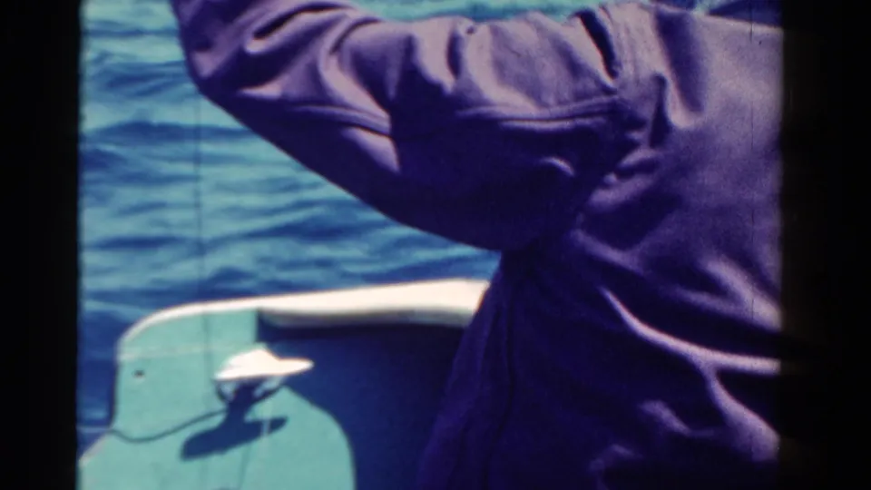 Home-movie frame from 1958 Ontario showing the fisherman from behind beside the green boat edge and rippling water.