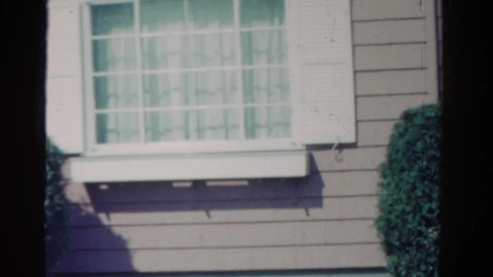 1958 Ontario home-movie frame showing a curtained house window, white shutters, clapboard siding, and a trimmed shrub.