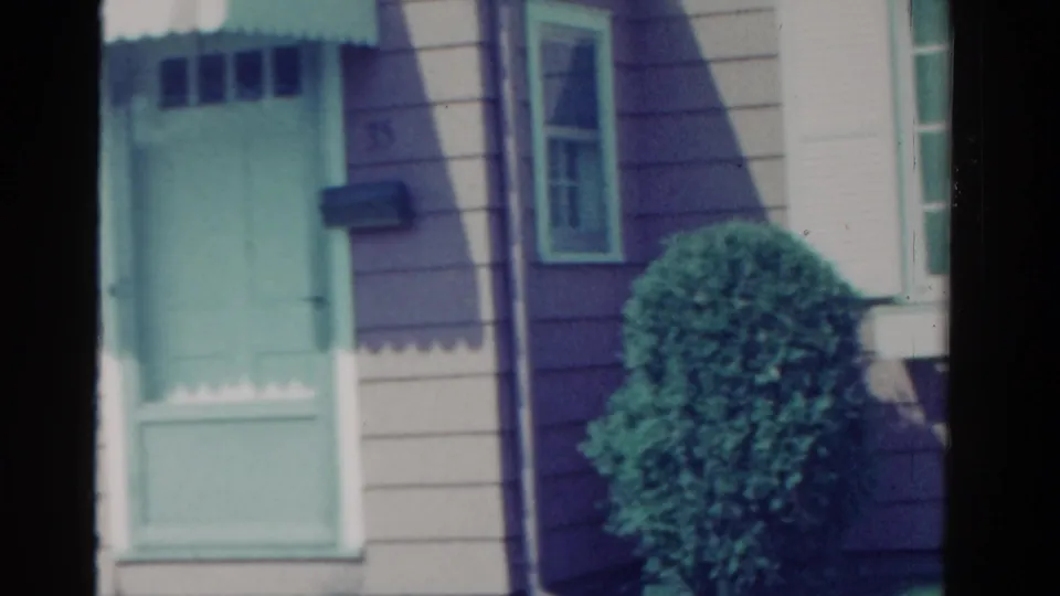 1958 Ontario house exterior with a green front door, wall mailbox, siding, window edge, and shrub during a handheld pan.
